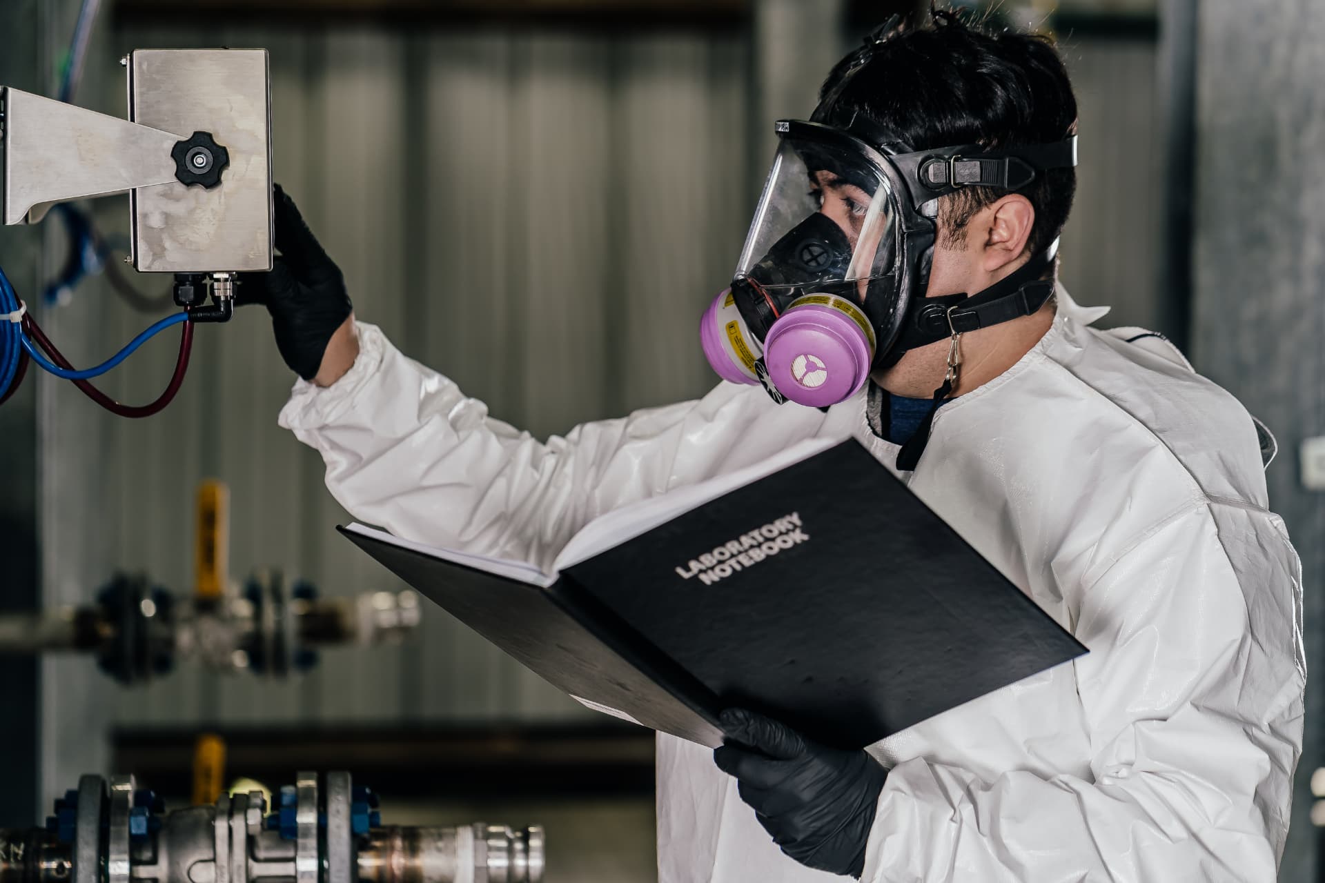 Element Chemicals technician in full-face respirator reviewing a laboratory notebook beside corrosion-inhibitor testing equipment